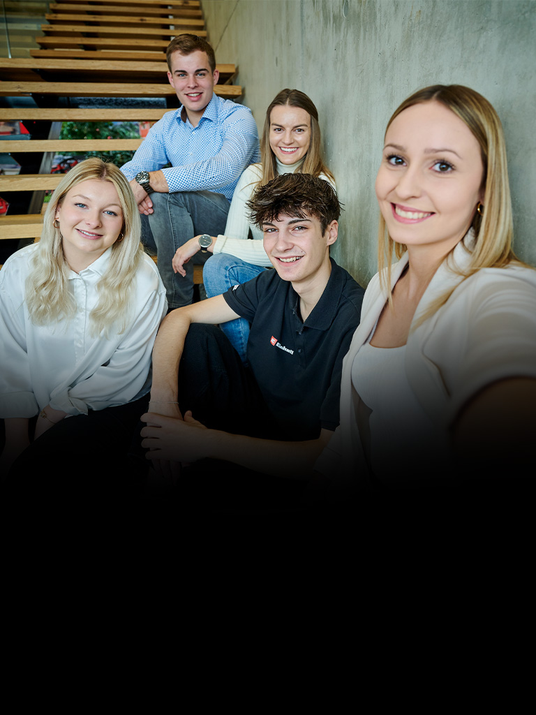 Five young adults sit on stairs taking a selfie, with part of the background in shadow.