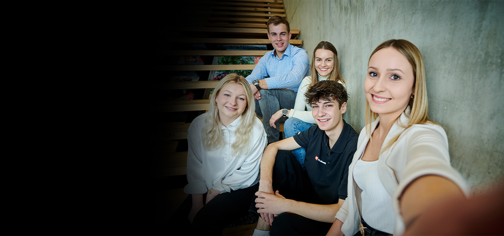 Five young adults sit on stairs taking a selfie, with part of the background in shadow.
