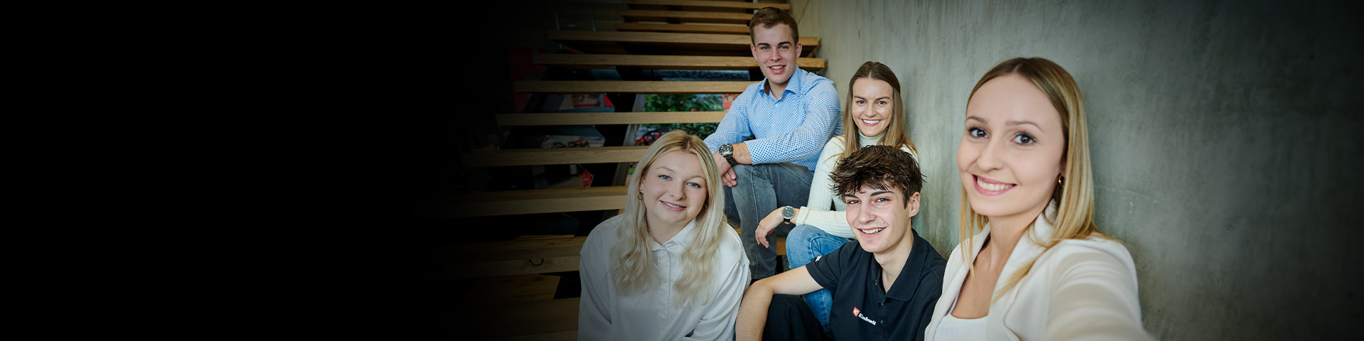 Five young adults sit on stairs taking a selfie, with part of the background in shadow.
