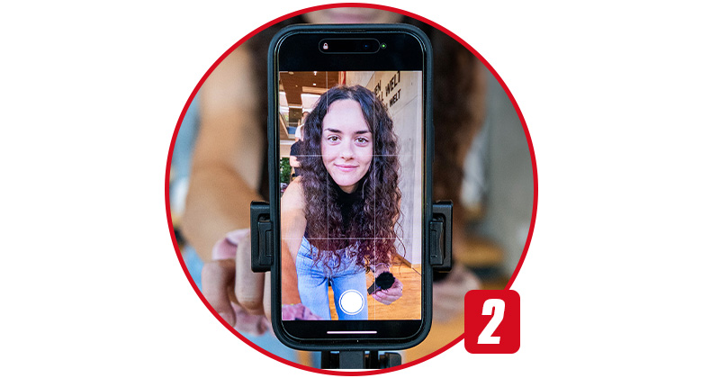 A woman with curly hair takes a selfie using a smartphone mounted on a holder.