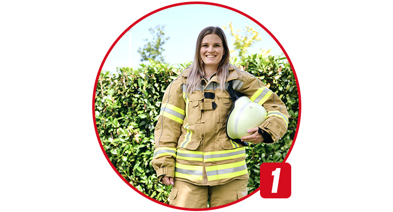 A young woman in full firefighter protective gear holds a firefighter helmet in her hand.