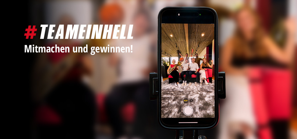 Auf einem Smartphone ist ein Gruppenfoto von sechs Personen mit Sportgeräten und Instrumenten zu sehen. Links steht: "#TEAMEINHELL Mitmachen und gewinnen!"